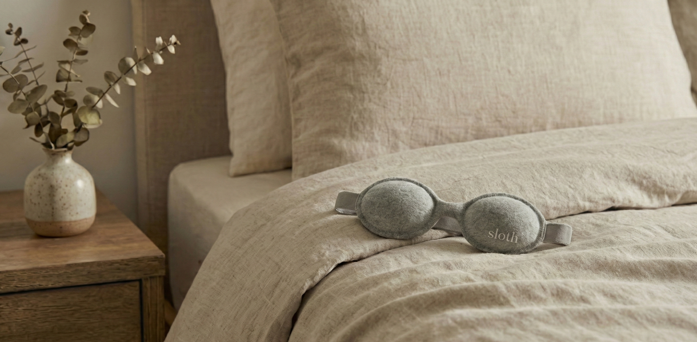 goggle style sleep mask on beige bedding with a vase of eucalyptus on a wooden nightstand.